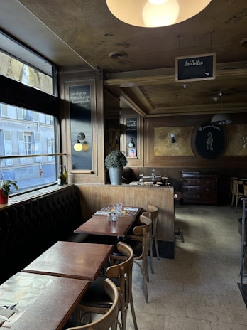A cozy cafe interior with wooden tables and chairs, decorated with potted plants and framed signs on the walls. The lighting is warm, and a large window allows natural light to illuminate the space. The overall ambiance is intimate and inviting.