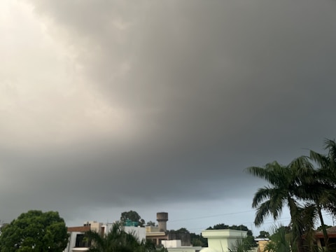 Dark storm clouds gather over a suburban neighborhood with homes and lush green trees, including palm trees, in the foreground.
