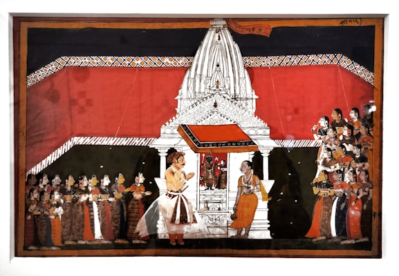 A detailed Indian painting showcases a traditional scene with a large white temple structure in the center. The temple is adorned with intricate designs and has a vivid red backdrop. In front of the temple, two figures are prominently positioned, one of whom appears to be in conversation with the other. To the left and right, groups of people are attentively gathered, wearing colorful traditional attire, indicating a ceremonial or religious gathering.