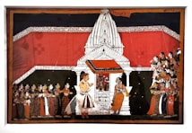 A detailed Indian painting showcases a traditional scene with a large white temple structure in the center. The temple is adorned with intricate designs and has a vivid red backdrop. In front of the temple, two figures are prominently positioned, one of whom appears to be in conversation with the other. To the left and right, groups of people are attentively gathered, wearing colorful traditional attire, indicating a ceremonial or religious gathering.