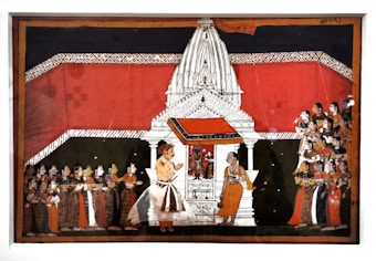 A detailed Indian painting showcases a traditional scene with a large white temple structure in the center. The temple is adorned with intricate designs and has a vivid red backdrop. In front of the temple, two figures are prominently positioned, one of whom appears to be in conversation with the other. To the left and right, groups of people are attentively gathered, wearing colorful traditional attire, indicating a ceremonial or religious gathering.