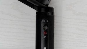 A close-up view of a black electronic device with buttons and switches on its surface. The device features a small joystick, a power button, and a mode switch with labels such as PF, L, F, and POV. The background is a light-colored surface with a subtle texture.