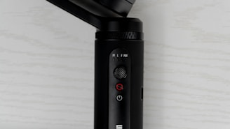 A close-up view of a black electronic device with buttons and switches on its surface. The device features a small joystick, a power button, and a mode switch with labels such as PF, L, F, and POV. The background is a light-colored surface with a subtle texture.