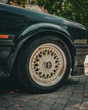Close-up view of a car's wheel and tire, with a stylish BBS rim and Hankook tire. The car's wheel arch and part of the side panel are visible, with reflections on the car's glossy dark paint. The car is parked on a cobblestone surface, with some leaves scattered around. In the background, there is greenery and the roof of a house partially visible.