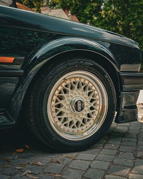 Close-up view of a car's wheel and tire, with a stylish BBS rim and Hankook tire. The car's wheel arch and part of the side panel are visible, with reflections on the car's glossy dark paint. The car is parked on a cobblestone surface, with some leaves scattered around. In the background, there is greenery and the roof of a house partially visible.