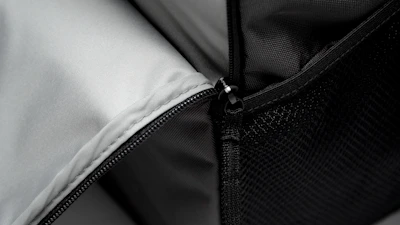A close-up view of a zipper on a fabric item. The zipper is partially opened, revealing a light gray inner lining. The outer fabric appears dark and textured, possibly from a bag or backpack.