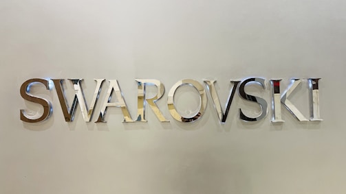 Metallic letters spelling 'SWAROVSKI' are mounted on a smooth, light-colored wall. The letters are reflective and have a shiny, polished finish.