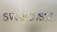 Metallic letters spelling 'SWAROVSKI' are mounted on a smooth, light-colored wall. The letters are reflective and have a shiny, polished finish.
