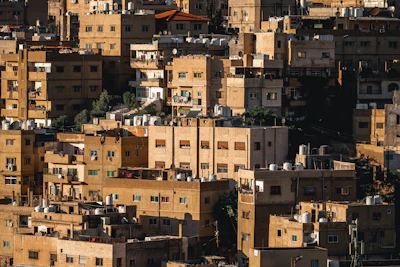 An urban landscape featuring densely packed apartment buildings with flat rooftops. The architecture is a mix of beige and light brown tones, with some buildings showing signs of wear. Various water tanks and satellite dishes are visible on the rooftops, and there is occasional greenery interspersed between the structures.