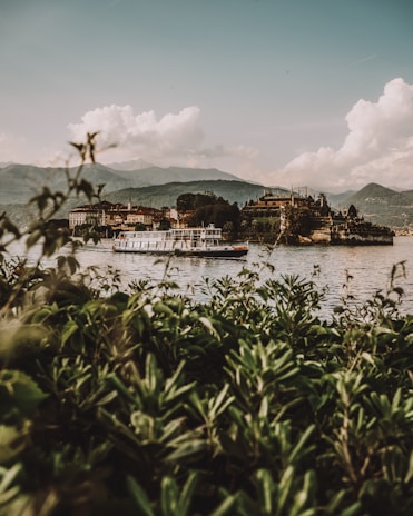 A serene landscape features a large river bordered by lush greenery in the foreground. A ferryboat glides across the water, with an intricate castle and quaint buildings nestled among trees on the opposite shore. Majestic mountains rise in the background under a sky filled with fluffy clouds.