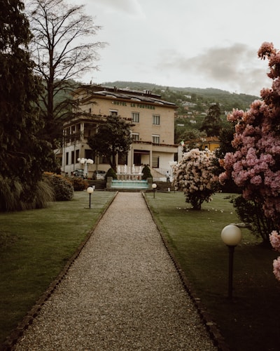 A gravel pathway leads through a well-maintained garden with neatly trimmed grass, flanked by trees and blossoming bushes with pink flowers. At the end of the path sits a large, elegant building with an inscription 'Hotel la Fontana'. The building has multiple floors and a classical architecture style, surrounded by a picturesque mountainous landscape in the distance. Soft lighting from round lampposts adds to the serene atmosphere.