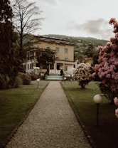 A gravel pathway leads through a well-maintained garden with neatly trimmed grass, flanked by trees and blossoming bushes with pink flowers. At the end of the path sits a large, elegant building with an inscription 'Hotel la Fontana'. The building has multiple floors and a classical architecture style, surrounded by a picturesque mountainous landscape in the distance. Soft lighting from round lampposts adds to the serene atmosphere.