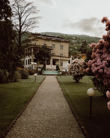 A gravel pathway leads through a well-maintained garden with neatly trimmed grass, flanked by trees and blossoming bushes with pink flowers. At the end of the path sits a large, elegant building with an inscription 'Hotel la Fontana'. The building has multiple floors and a classical architecture style, surrounded by a picturesque mountainous landscape in the distance. Soft lighting from round lampposts adds to the serene atmosphere.