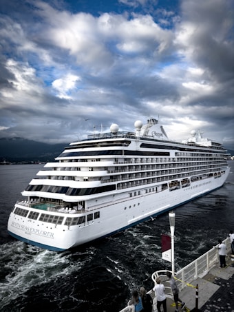 A large white cruise ship named Seven Seas Explorer is sailing through a body of water. The sky is filled with dramatic clouds, and people are observing the ship from a dock.