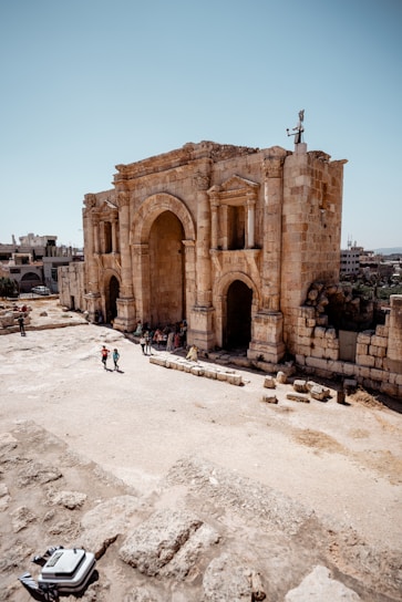 An ancient stone structure with large arches, likely a historical or archaeological site. Several people are gathered near the building, suggesting it might be a tourist attraction. The ground around is sandy with scattered stones, under a clear blue sky.