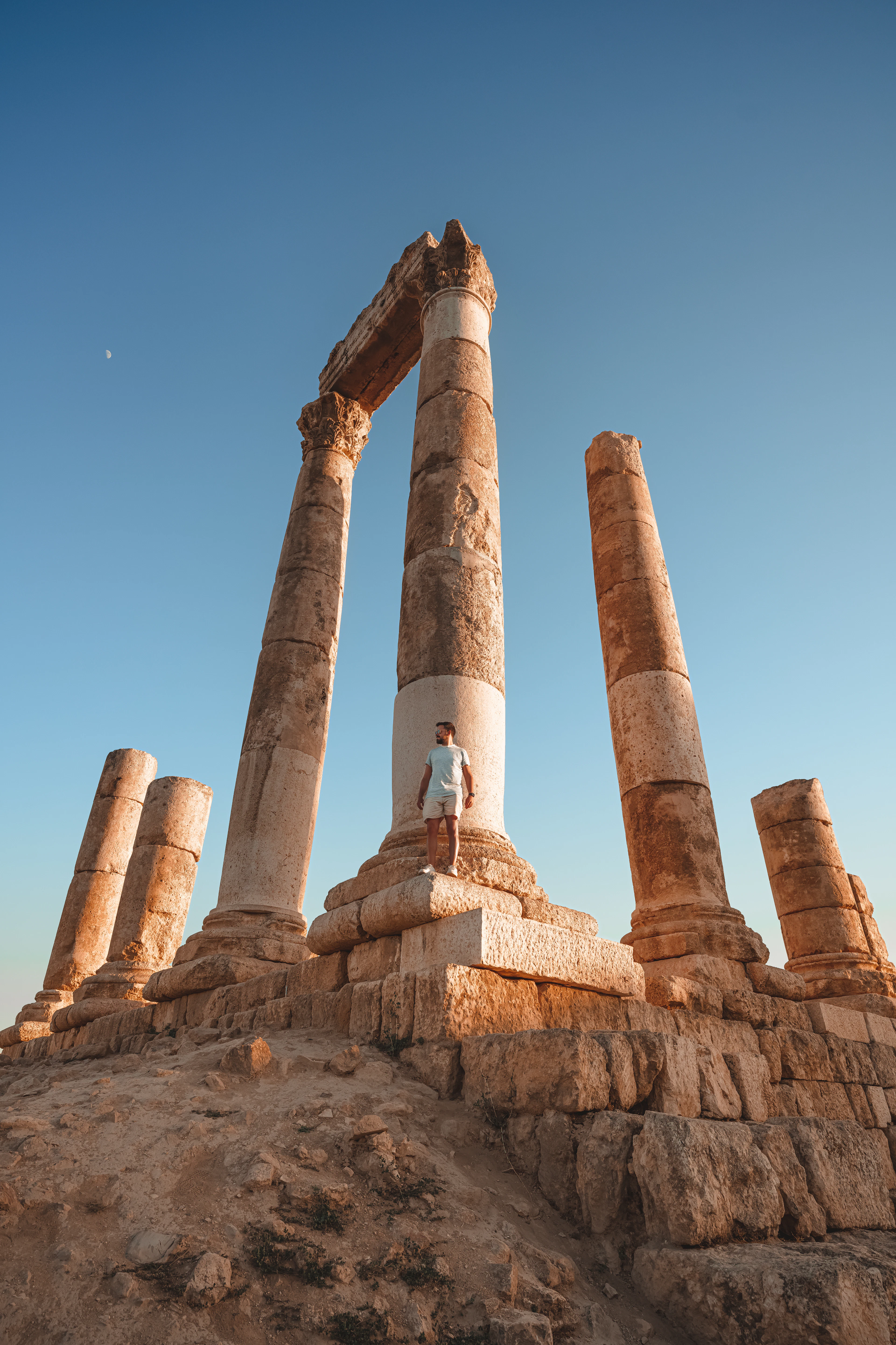 The towering columns of the Temple of Hercules on the Amman Citadel at golden hour, a lone visitor standing on the podium at their base for scale against a deep blue sky.