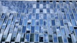 A building facade featuring a grid of glass windows that reflect the blue sky and white clouds, creating a pattern of alternating light and dark tones. The reflection captures partial images of surrounding structures, adding a dynamic and modern architectural element.
