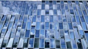 A building facade featuring a grid of glass windows that reflect the blue sky and white clouds, creating a pattern of alternating light and dark tones. The reflection captures partial images of surrounding structures, adding a dynamic and modern architectural element.