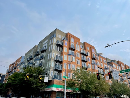 A multi-story residential building with modern architecture, featuring a combination of gray and orange facades. The building has numerous balconies and large windows. There are traffic lights and street signs in the foreground, with trees lining the street.