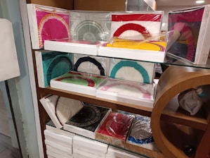 Multiple boxes containing colorful paper fans are displayed neatly on glass shelves with a wooden frame. The fans are packaged in plastic, and the boxes are stacked in an organized manner. The fans come in various colors such as red, yellow, green, blue, pink, black, and white. Some of the boxes have fans with dual color combinations.