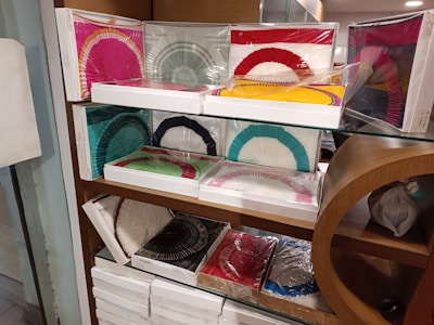 Multiple boxes containing colorful paper fans are displayed neatly on glass shelves with a wooden frame. The fans are packaged in plastic, and the boxes are stacked in an organized manner. The fans come in various colors such as red, yellow, green, blue, pink, black, and white. Some of the boxes have fans with dual color combinations.