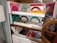 Multiple boxes containing colorful paper fans are displayed neatly on glass shelves with a wooden frame. The fans are packaged in plastic, and the boxes are stacked in an organized manner. The fans come in various colors such as red, yellow, green, blue, pink, black, and white. Some of the boxes have fans with dual color combinations.