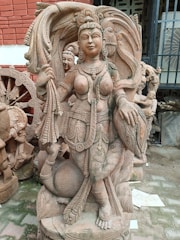 A detailed stone sculpture of an intricately carved deity with elaborate adornments and a divine posture, surrounded by other figures and decorative elements. The craftsmanship highlights traditional attire and symbolic gestures, conveying a sense of grace and spirituality.