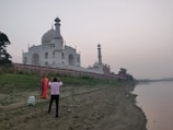 A person in a pink shirt is taking a photo of a person in traditional attire near the Taj Mahal, which stands prominently in the background. The monument is surrounded by lush greenery and adjacent to a calm river. The sky appears overcast.