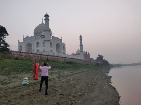 A person in a pink shirt is taking a photo of a person in traditional attire near the Taj Mahal, which stands prominently in the background. The monument is surrounded by lush greenery and adjacent to a calm river. The sky appears overcast.
