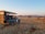 A safari vehicle with an open roof is parked on a dry, grassy landscape. Several people are seated in the vehicle, observing the surroundings, possibly on a wildlife expedition. The area is filled with shrubs and sparse trees under a clear sky, with distant mountains visible on the horizon.