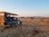 A safari vehicle with an open roof is parked on a dry, grassy landscape. Several people are seated in the vehicle, observing the surroundings, possibly on a wildlife expedition. The area is filled with shrubs and sparse trees under a clear sky, with distant mountains visible on the horizon.