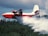 A large red and white aircraft is releasing a stream of water over a dense forest. The action suggests that the plane is involved in aerial firefighting. The sky is cloudy, adding to the dramatic effect of the scene.