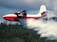 A large red and white aircraft is releasing a stream of water over a dense forest. The action suggests that the plane is involved in aerial firefighting. The sky is cloudy, adding to the dramatic effect of the scene.