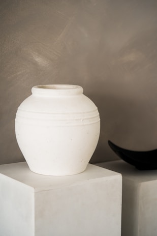 A white ceramic pot with a smooth surface sits on a white pedestal. The background is a muted gray wall, providing a minimalist and modern aesthetic. To the right, a black object, possibly a decorative bowl or sculpture, partially appears.