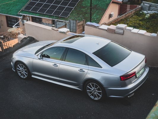 A sleek silver sedan parked on a sloped driveway next to a house with green roofing and solar panels installed. The car features modern design elements, such as alloy wheels and tinted windows. The background includes rising staircases and stacked tiles on the wall.