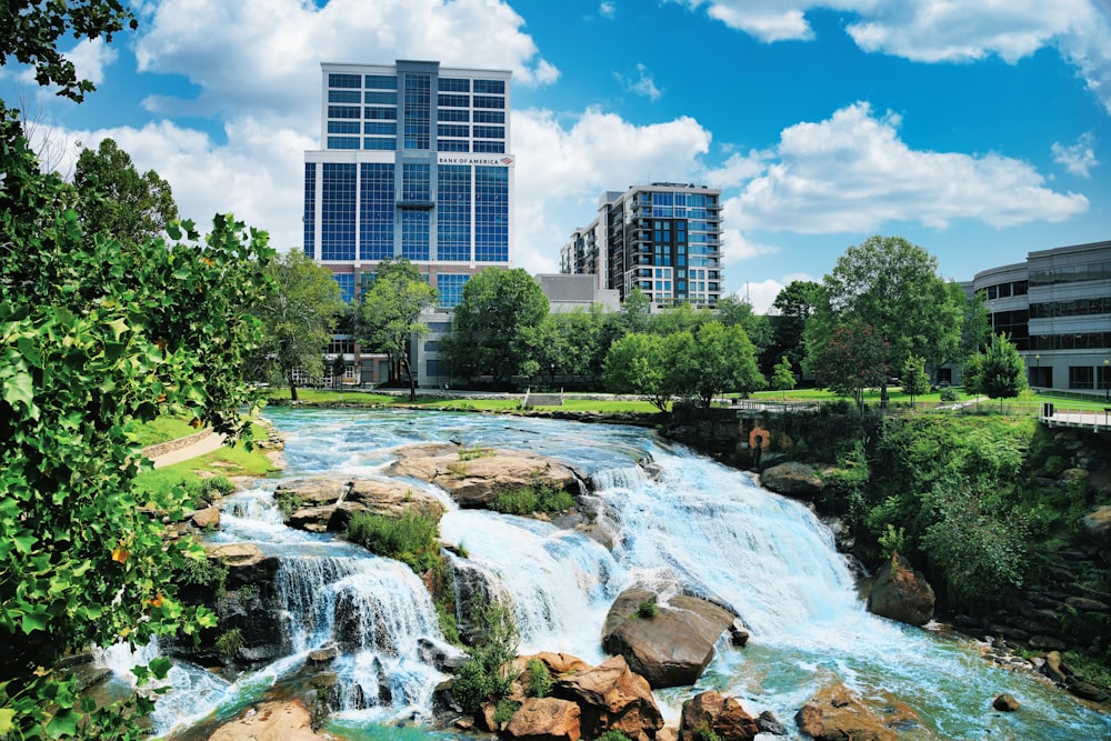 a waterfall in the middle of a park with buildings in the background