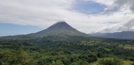 A large volcano rises majestically in the distance, surrounded by dense, green forest. The summit of the volcano is lightly capped with white clouds, and the sky above is partly cloudy with patches of blue visible.