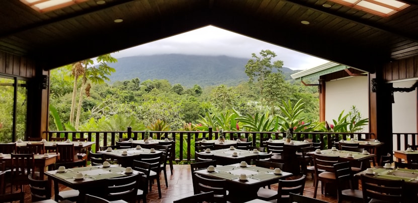 The image depicts an open-air dining area with several wooden tables and chairs set with dishes and cutlery, overlooking a lush, green forest and distant mountains partially obscured by clouds.
