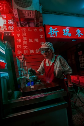 A street food vendor is making a dish in a dimly lit setting. The vendor is wearing a white shirt with an apron and a cap, standing behind a counter with cooking utensils. Bright red and neon lighting from signage illuminates the area, casting a vibrant glow against the vendor and the cooking setup.