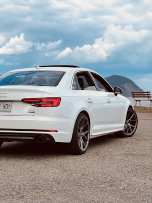 A white sedan is parked on a paved surface with a scenic backdrop of mountains and a partly cloudy sky. The rear of the car displays a Quebec license plate and features like the taillights and alloy wheels are visible. A wooden bench is situated nearby, suggesting a lookout point or rest area.