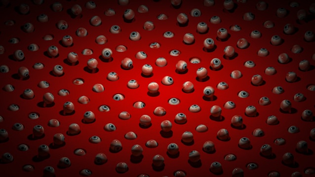 An array of realistic artificial eyeballs spread across a red surface, each eyeball featuring detailed irises and pupils. The eyeballs are positioned at various angles, casting small shadows on the red background.