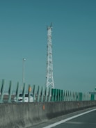 A tall communication tower stands alongside a highway with green protective barriers in the foreground. A white car is visible on the road, partially obscured by the barriers. The sky is clear, creating a calm backdrop.