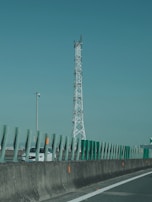 A tall communication tower stands alongside a highway with green protective barriers in the foreground. A white car is visible on the road, partially obscured by the barriers. The sky is clear, creating a calm backdrop.