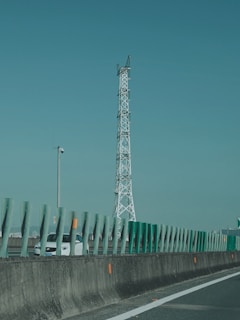 A tall communication tower stands alongside a highway with green protective barriers in the foreground. A white car is visible on the road, partially obscured by the barriers. The sky is clear, creating a calm backdrop.