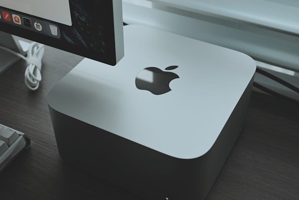 A sleek, silver-colored computer device with an Apple logo is placed on a dark wooden surface. Behind the device, part of a monitor is visible, displaying a row of application icons. A white keyboard cable is slightly visible, adding a modern and tech-focused feel to the setup.