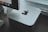 A sleek, silver-colored computer device with an Apple logo is placed on a dark wooden surface. Behind the device, part of a monitor is visible, displaying a row of application icons. A white keyboard cable is slightly visible, adding a modern and tech-focused feel to the setup.