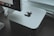 A sleek, silver-colored computer device with an Apple logo is placed on a dark wooden surface. Behind the device, part of a monitor is visible, displaying a row of application icons. A white keyboard cable is slightly visible, adding a modern and tech-focused feel to the setup.