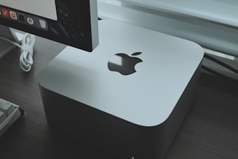 A sleek, silver-colored computer device with an Apple logo is placed on a dark wooden surface. Behind the device, part of a monitor is visible, displaying a row of application icons. A white keyboard cable is slightly visible, adding a modern and tech-focused feel to the setup.