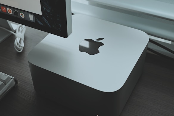 A sleek, silver-colored computer device with an Apple logo is placed on a dark wooden surface. Behind the device, part of a monitor is visible, displaying a row of application icons. A white keyboard cable is slightly visible, adding a modern and tech-focused feel to the setup.