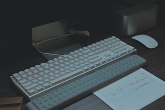 A modern workspace setup featuring a white mechanical keyboard with a wrist rest, an Apple Magic Mouse, and a note with handwritten text on lined paper. There is a mini PC or docking station nearby, along with a monitor. The lighting is dim, creating a moody atmosphere.